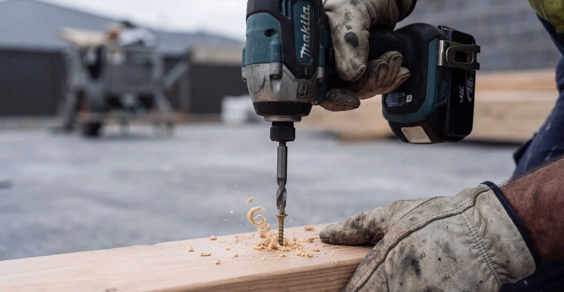 A gloved hand driving a screw into timber, shavings coming off the bit.